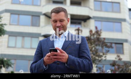Felice bello e sicuro ragazzo in completo che digita al telefono cellulare e sorride di buon umore. Primo piano del gioioso uomo d'affari che invia messaggi di testo sullo smartphone Foto Stock