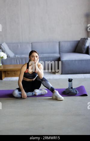 Soddisfatta blogger europea bella e giovane bionda che si riposa dopo l'allenamento a casa con l'acqua, donna prende una pausa sul tappeto nella palestra domestica. Vita sana Foto Stock