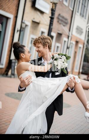 amore in città, felice giovane sposo che tiene la sposa afroamericana con fiori in strada Foto Stock