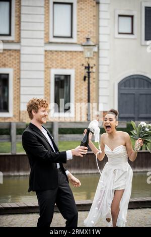 eccitata sposa afroamericana vicino allo sposo con champagne versato dalla bottiglia, festeggiamenti all'aperto Foto Stock