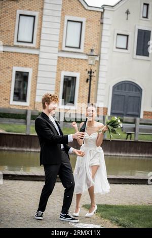 sposo con champagne versato dalla bottiglia vicino alla felicissima sposa afroamericana, festeggiamenti all'aperto Foto Stock