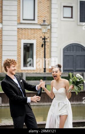 matrimonio in città, sposo rosso con champagne versato dalla bottiglia vicino a spose afroamericane urlanti Foto Stock