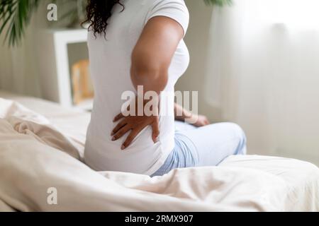 Giovane donna nera avendo indietro Ache mentre seduta su letto a casa Foto Stock