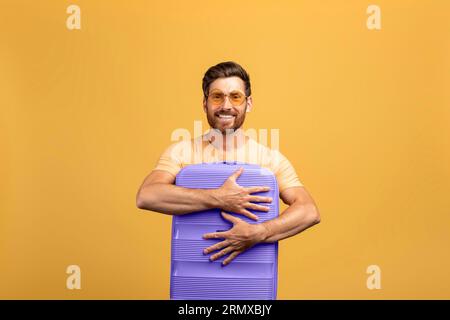Foto di un uomo europeo di mezza età che abbraccia la sua nuova valigia per il viaggio, in piedi su sfondo giallo Foto Stock