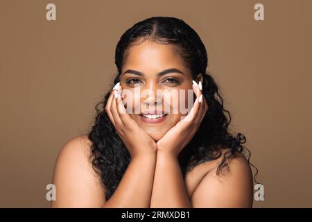 Concetto di cura del viso. Bella donna brasiliana positiva con capelli ricci che tocca il viso con una pelle perfettamente liscia Foto Stock