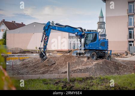 Escavatore o retroescavatore in un cantiere in città. Scavatrice blu appoggiata su un cumulo di sporcizia o ghiaia mentre si crea la piastra di base per un edificio. Foto Stock