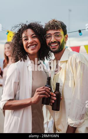 Ritratto verticale di una giovane coppia multirazziale che sorride, ride e guarda la macchina fotografica con le bottiglie di birra. Due persone che guardano davanti a una festa con i loro migliori amici. Foto di alta qualità Foto Stock