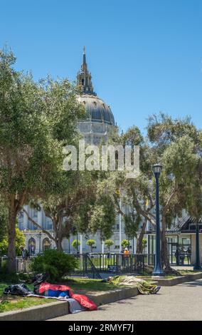 San Francisco, CA, USA - July 12, 2023: Homeless people sleep on Civic Center Plaza in front of City Hall. Its dome peeks over tree foliage under blue Stock Photo