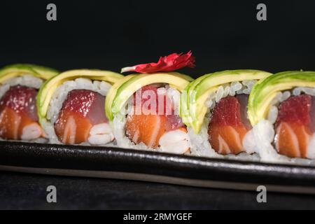 Deliziosi involtini giapponesi di sushi uramaki con fette di avocado in cima serviti su vassoio su superficie nera sfocata Foto Stock