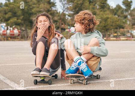 Deliziati i ragazzi e le ragazze che soffiano gomma da masticare mentre si siedono sugli skateboard in città e si divertono insieme durante il fine settimana Foto Stock