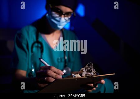 Basso angolo di medico femminile concentrato in uniforme e maschera protettiva seduto sulle scale con appunti e prendere appunti in foglio di storia medica Foto Stock