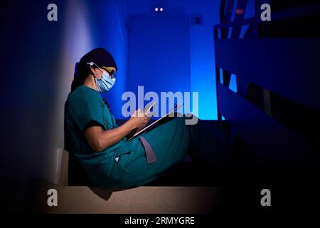 Vista laterale del medico femminile in maschera medica e abito seduto su scala e navigazione telefono cellulare in camera scura sotto illuminazione blu Foto Stock