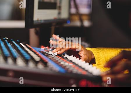 Una donna afroamericana di coltura irriconoscibile in maglione giallo con pannello di miscelazione e pulsanti di controllo mentre lavora in studio di trasmissione Foto Stock