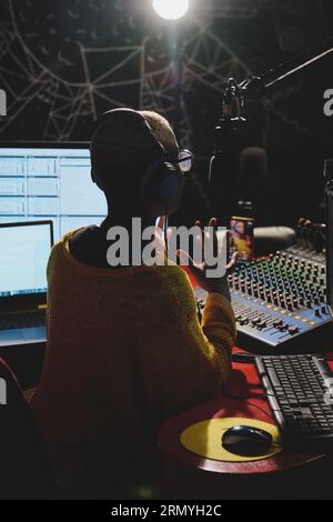 Vista posteriore di un'anonima conduttrice radiofonica etnica con le cuffie che registrano podcast mentre si siede al tavolo in uno studio di trasmissione al buio Foto Stock