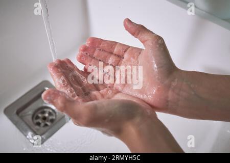 Mani di prodotto senza volto bambini lavarsi le mani con sapone schiumoso nel lavandino con flusso d'acqua durante la routine igienica in bagno Foto Stock