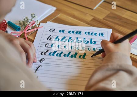 Dall'alto di un artista irriconoscibile che impara l'arte di scrivere disegnare su un foglio di carta allineato con schemi di scrittura a lettere posizionati su un tavolo di legno utilizzando bl Foto Stock