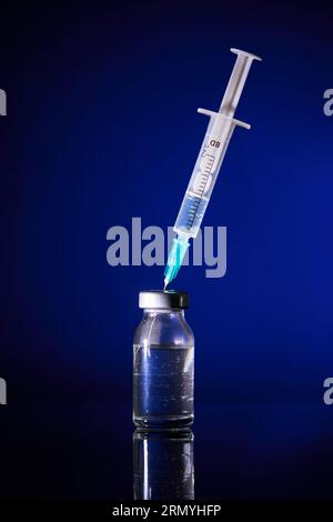 Siringa medica con ago inserito nel flacone trasparente di vaccino posto su sfondo blu in studio Foto Stock