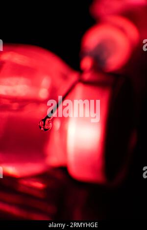 Concentrarsi sulla goccia di vaccino alla punta dell'ago posta su una fiala di vetro alla luce rossa su sfondo nero Foto Stock