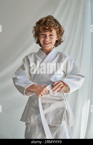 Ragazzo sorridente dai capelli ricci in kimono bianco in piedi vicino alla tenda e alla cintura mentre guarda la fotocamera prima di allenarsi con il judo Foto Stock