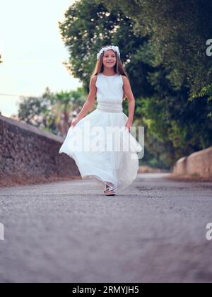 Tutta la lunghezza di Happy Girl con una corona bianca che regge l'orlo di un lungo abito da sole bianco mentre passeggi lungo la strada vicino agli alberi Foto Stock