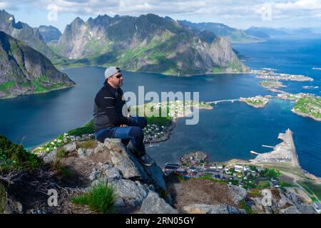 Rilassato e felice escursionista gode di una vista mozzafiato sulle cime di Reinebringen delle isole Lofoten, dei fiordi e della Norvegia Foto Stock