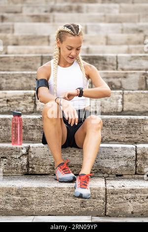 Runner donna che controlla i risultati su orologi intelligenti, seduto sulle scale, che prende il polso dopo aver fatto jogging e riposato, fascia da braccio con telefono cellulare. Foto Stock