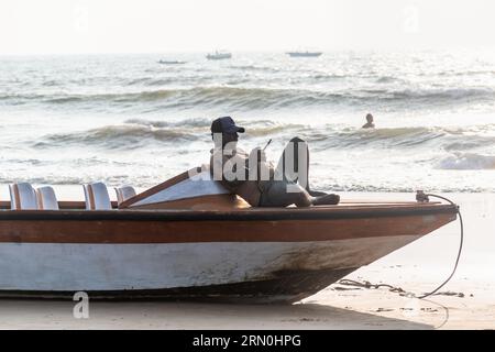 Calangute, Goa, India - January 2023: An Indian fisherman relaxing on a boat staring at his phone on a beach in Goa. Foto Stock