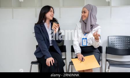 Due giovani candidate asiatiche amichevoli e felici si divertono a parlare davanti alla stanza per un colloquio di lavoro. Prima jobbers, internsh Foto Stock