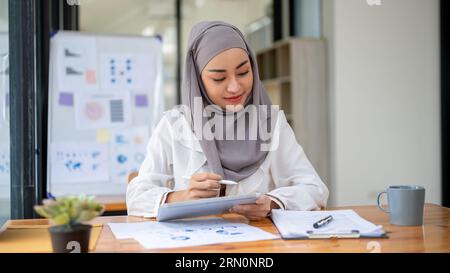 Un'attraente donna d'affari musulmana asiatica o una commercialista che indossa un hijab si concentra sui rapporti finanziari, lavora alla sua scrivania in ufficio. Foto Stock