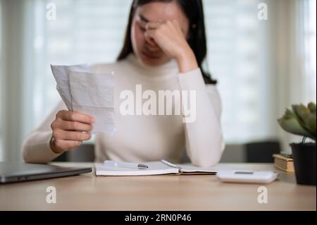 A stressed young Asian woman is holding invoices or bills, calculating her monthly household expenses, and worried about financial problems while sitt Foto Stock