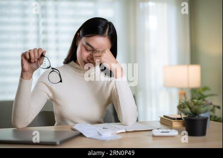 Una giovane donna asiatica, stressata, sta calcolando le spese mensili della sua famiglia, sentendosi frustrata, avendo mal di testa e preoccupata per i problemi finanziari Foto Stock
