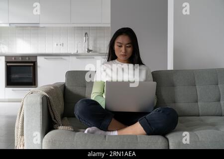 Una donna coreana professionista concentrata siede sul divano con un computer portatile e scrive testo in remoto Foto Stock