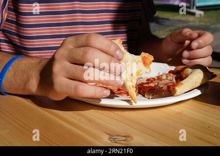 Ragazzo che mangia pizza con le mani nel ristorante all'aperto durante la giornata di sole. Ha una t-shirt rossa con cinturini e un bracciale in gomma blu sulla mano destra. Foto Stock