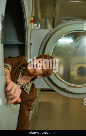 elegante rossa e un giovane barbuto che guarda la macchina fotografica dalla lavatrice nella lavanderia a gettoni Foto Stock