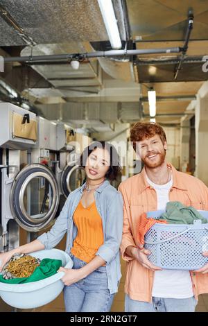 coppia interrazziale positiva che tiene cesti con vestiti e guarda la macchina fotografica nella lavanderia a gettoni Foto Stock