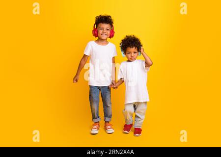 Full length photo of cheerful positive small boys wear white t-shirt walking hands listening music headphones isolated yellow color background Stock Photo