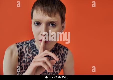 persona androgina con abito stampa leopardo che mostra un cartello di hush su sfondo arancione, stile queer Foto Stock