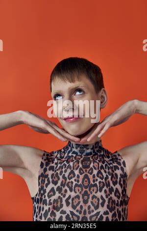 felice persona androgina sorridente e in posa su sfondo arancione, mani vicine al viso, stile queer Foto Stock