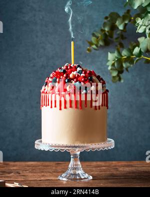 Torta di compleanno cremosa con gocce rosse e molte caramelle e frutti di bosco. In piedi su un elegante banco per dolci in vetro su un tavolo rustico in legno. Foto Stock