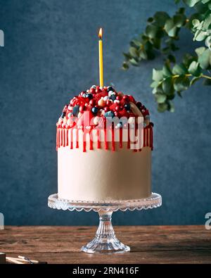 Torta di compleanno cremosa con gocce rosse e molte caramelle e frutti di bosco. In piedi su un elegante banco per dolci in vetro su un tavolo rustico in legno. Foto Stock