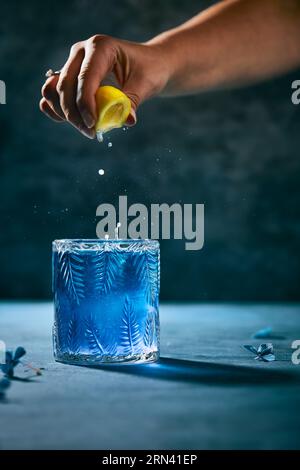 Cocktail blu scuro. Spremere a mano il succo di limone nel cocktail. Tavolo blu scuro e fiori su di esso. Sfondo nero. Immagine verticale dello stile di vita. Foto Stock