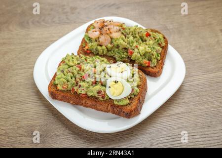 Fette di pane con guacamole saporito, uova e gamberi su tavola di legno, primo piano Foto Stock