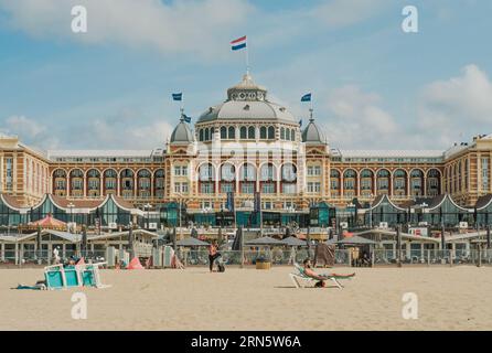 Grand Hotel Amrâth Kurhaus as seen from the front and centered, with restaurants and people lounging on the beach in front Stock Photo