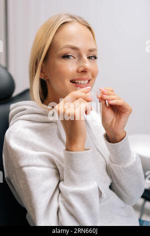 Ritratto verticale di una paziente bionda sorridente che regge un apparecchio invisibile e allineato seduto sulla sedia in una clinica odontoiatrica. Foto Stock