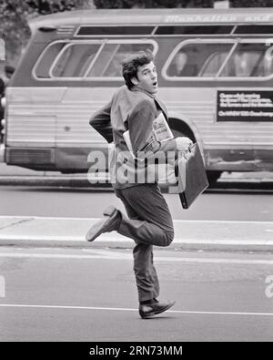 1970s YOUNG BUSINESSMAN RUNNING TO CATCH MORNING BUS WITH BRIEFCASE AND NEWSPAPER UNDER ARM  - s19149 HAR001 HARS CATCH FULL-LENGTH PERSONS MALES TRANSPORTATION EXPRESSIONS B&W LATE MORNING EYE CONTACT COMMUTE SUIT AND TIE SELLING MOTOR VEHICLE AND EXCITEMENT DIRECTION COMMUTING CONNECTION MOTION BLUR CITIES ATTACHE CASE BUSES SALESMEN TRANSIT YOUNG ADULT MAN BLACK AND WHITE CAUCASIAN ETHNICITY COMMUTER HAR001 HURRY MOTOR VEHICLES OLD FASHIONED Stock Photo