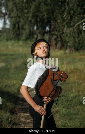 Ritratto di una ragazza - un fantino in un casco e una maglietta bianca, che abbraccia un cavallo marrone - un giocattolo su un bastone, guarda nella macchina fotografica. Alberi verdi nel ba Foto Stock