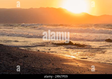 Waves hitting an idyllic beach in Greece. Summer day with rough sea. In the background the Sun sets behind the mountains Stock Photo