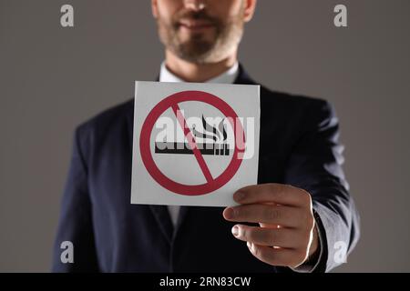 Uomo che tiene una carta con un cartello di divieto di fumo su sfondo grigio, primo piano Foto Stock