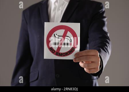 Uomo che tiene una carta con un cartello di divieto di fumo su sfondo grigio, primo piano Foto Stock