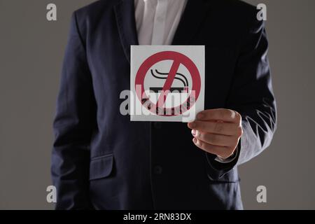 Uomo che tiene una carta con un cartello di divieto di fumo su sfondo grigio, primo piano Foto Stock